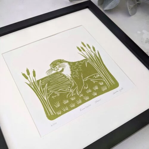 Linocut print of an otter on a riverbank framed by bullrushes in green - rose and hen