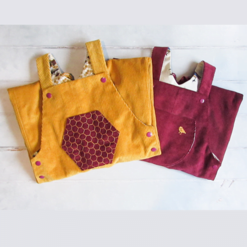 An image showing a pair of gold corduroy dungarees, with a hand embroidered honeycomb pocket, folded on top of a pair of dark red corduroy dungarres with a gold birth embroidered in the top right of the pocket.