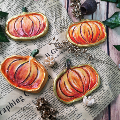 Four clay, hand painted pumpkin trinket trays on newscript with seed pods and dried flora scattered around them.