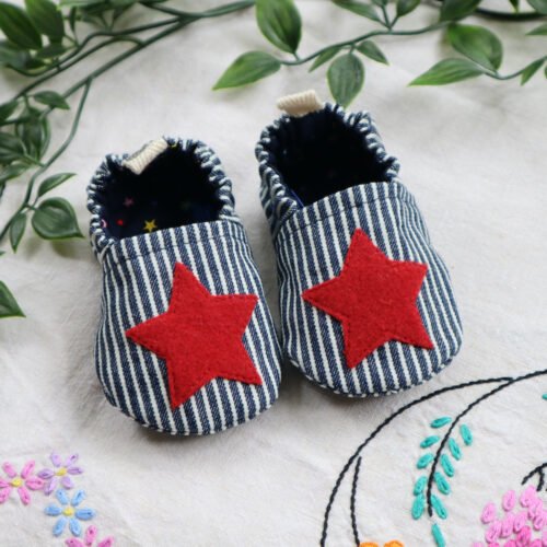 A pair of soft baby shoes made in a striped blue and white denim and red stars appliquéd onto the uppers