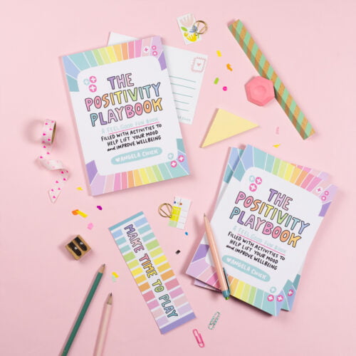 A photograph of a book laid out on a table with some stationery surrounding it. The book has a rainbow cover and is called The Positivity Playbook