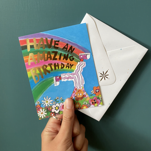 Hand holding a birthday card designed in Canada. The card is a small kid doing a cartwheel with a rainbow following them, in the rainbow it reads have an amazing birthday.