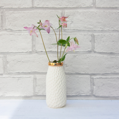 Image of small porcelain white milk bottle vase with a shiny gold top rim, with pink flowers, against a white brick wall on a white table.