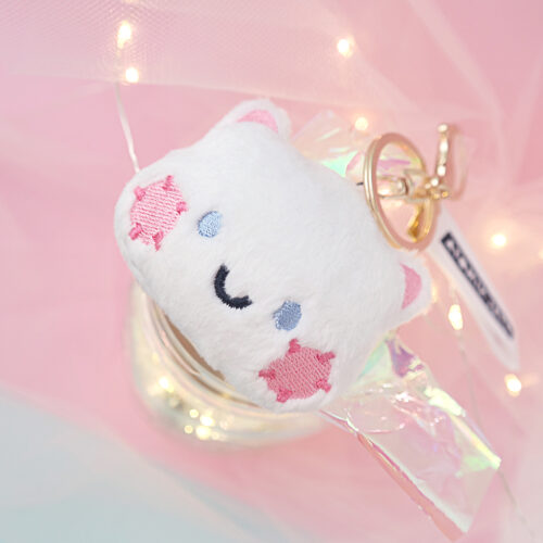 White plushie cat sitting on pink background with tulle and lights. Cat has blue eyes and pink cheeks and a smile. With a Gold lobster clasp